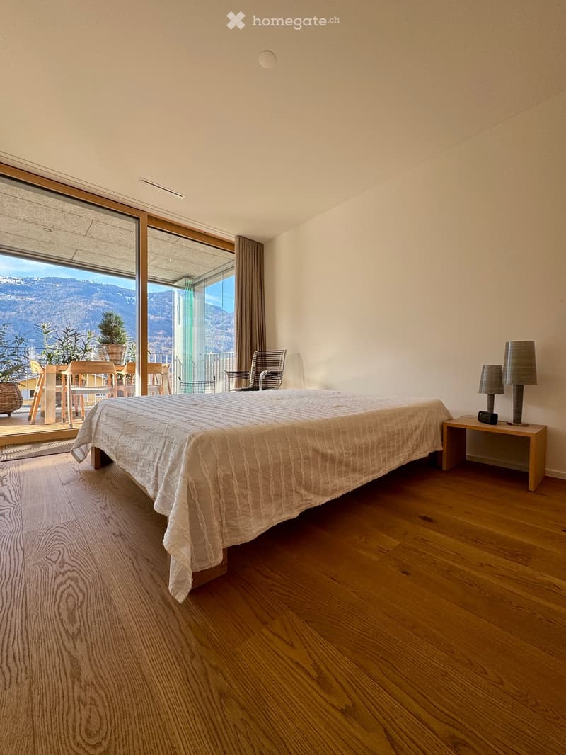 Large bedroom, hardwood floor, queen-size bed, white bedding, nightstands with lamps, large sliding glass doors to a balcony, table and chairs with potted plants on the balcony, scenic mountain view