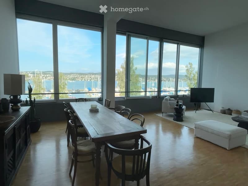 Bright, open living and dining area with large windows offering a scenic view of a harbor and cityscape. The space features a wooden dining table with chairs, a white sofa with an ottoman, a floor lamp, and a television on a stand.
