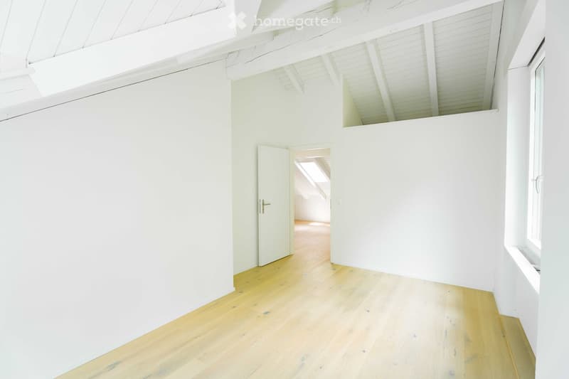 Empty room with white walls, wooden floor, and a door leading to another space. The ceiling has exposed beams and a skylight.