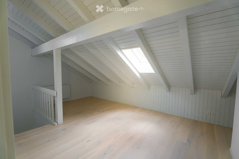 An empty attic space with wooden flooring, white wooden beams, and exposed ceiling beams. The room has white walls and a small railing along one side.