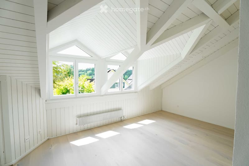 Bright, empty attic room with wooden walls, ceiling beams, and large windows letting in natural light. The floor is light wooden, and there is a radiator on one wall.