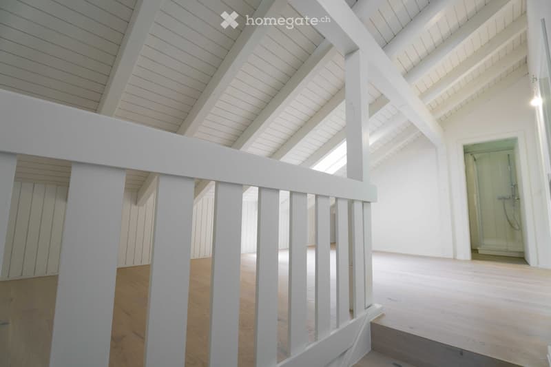An empty, open-plan space with wooden flooring and white wooden railings, featuring a visible bathroom area with a glass shower enclosure.