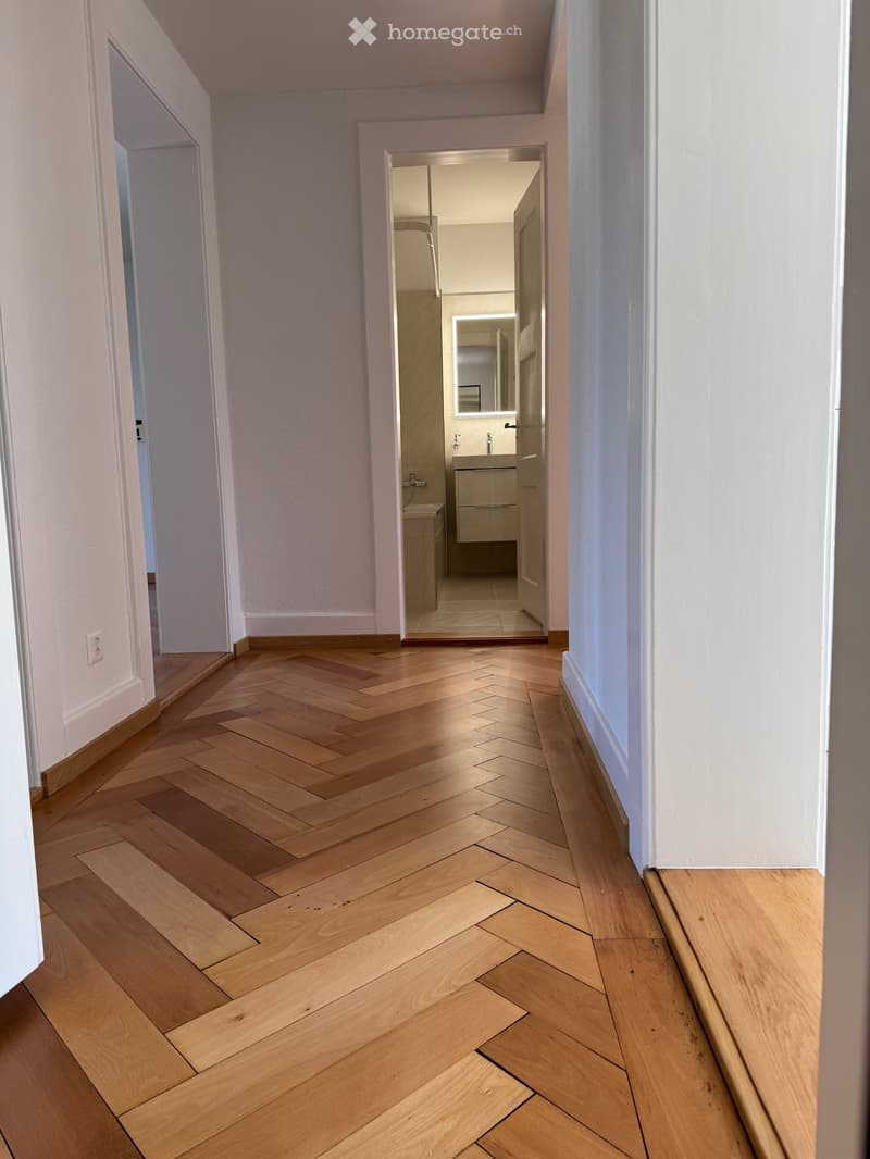 Empty hallway with wooden parquet flooring, white walls, and a bathroom visible through an open door.