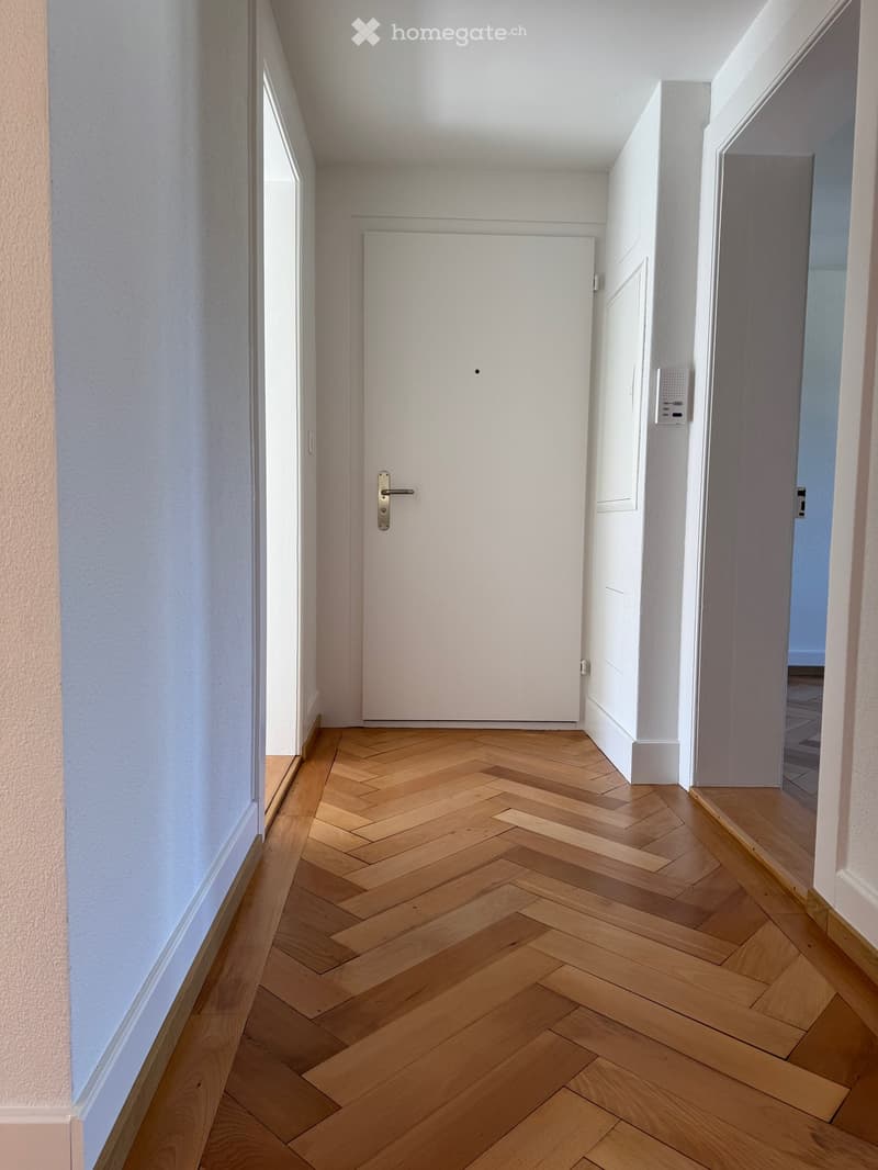 Empty hallway with wooden parquet flooring, white walls, white door, and a small window