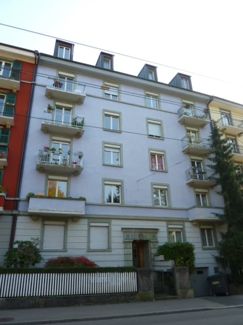 white building, 4 levels, multiple balconies, large windows, trees, trash bin