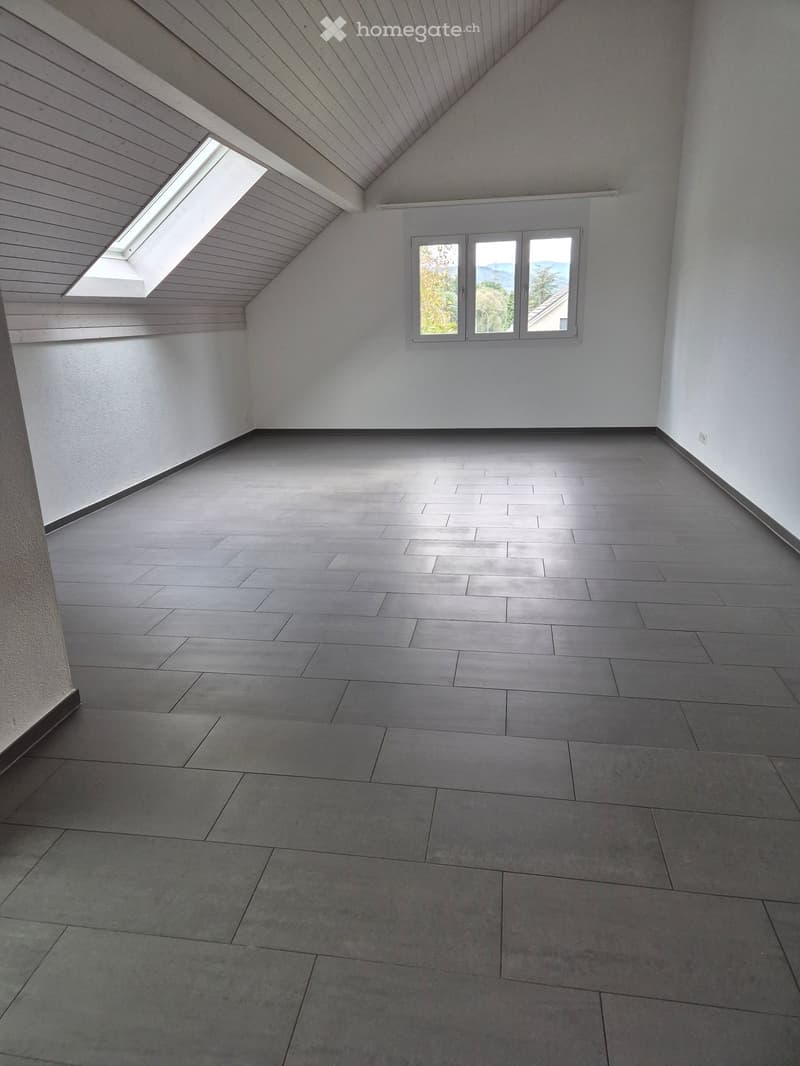 Empty room with grey tiled floor, white walls, and a skylight window, featuring a slanted wooden ceiling and a large window with a view.