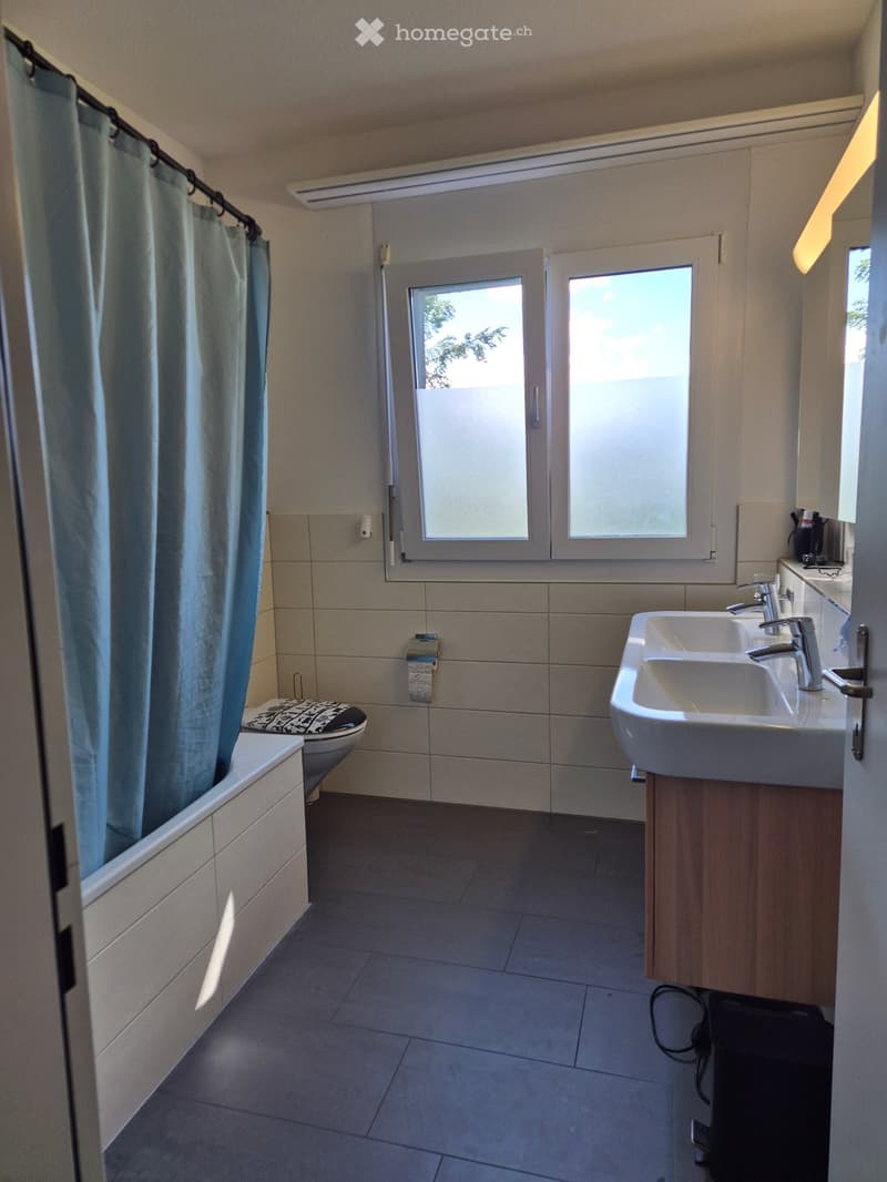 Modern bathroom with a bathtub, toilet, double sink, and large windows. Light-colored tiles and wooden cabinetry.