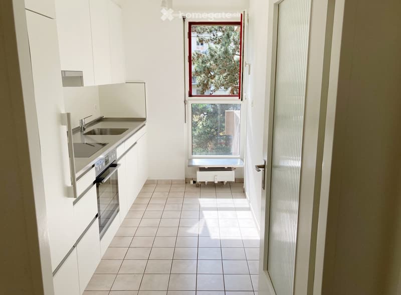 Modern, minimalist kitchen with white cabinets, a sink, and an oven. The space is bright with tiled flooring and large windows offering a view of greenery.