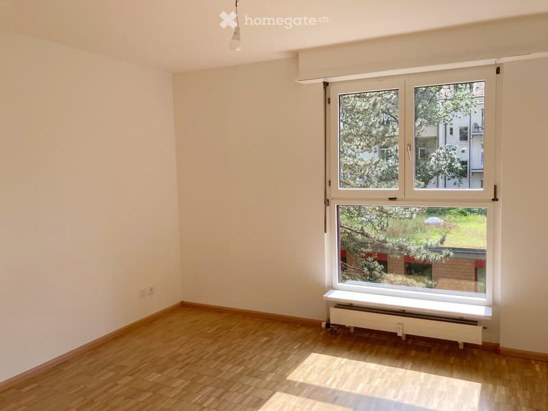 Empty room with wooden flooring, white walls, large window with blinds, radiator, and visible electrical outlets.