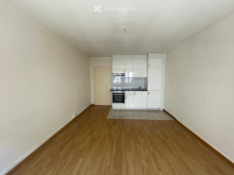 Empty room with light wooden flooring, white walls, and a small white kitchen area with cabinets, sink, and stove.
