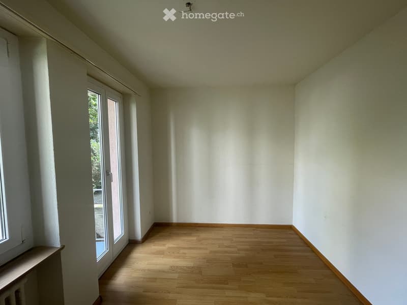 Empty room with wooden flooring, white walls, and large windows with sliding doors leading to an outdoor area.