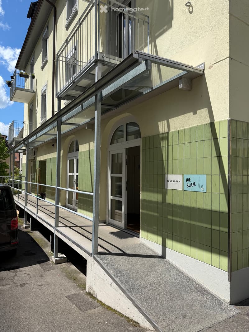 Exterior view of a multi-story building with a covered entrance, glass doors, and a ramp leading to a balcony area with metal railings.