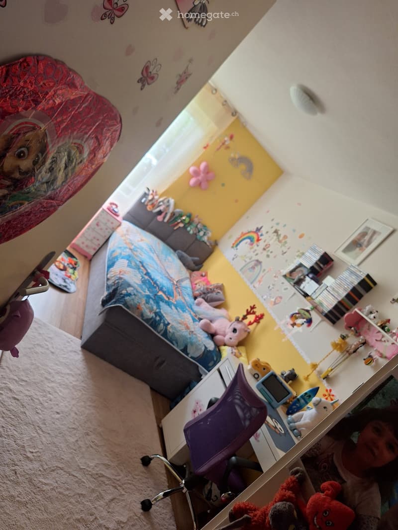 A child's bedroom with a bed, desk, chair, and various toys and decorations, including a colorful wall with stickers and a window letting in natural light.