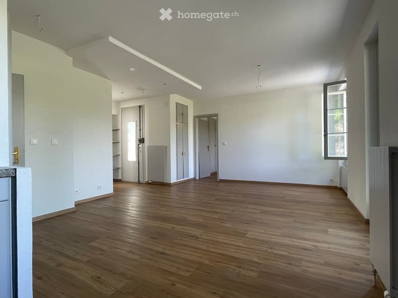 Empty room with wooden flooring, white walls, and large windows allowing natural light. The space features a door leading to another room and built-in cabinets.