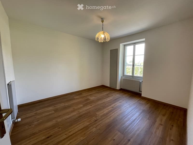 Empty room with wooden flooring, white walls, a window with a radiator below, and a hanging light fixture.
