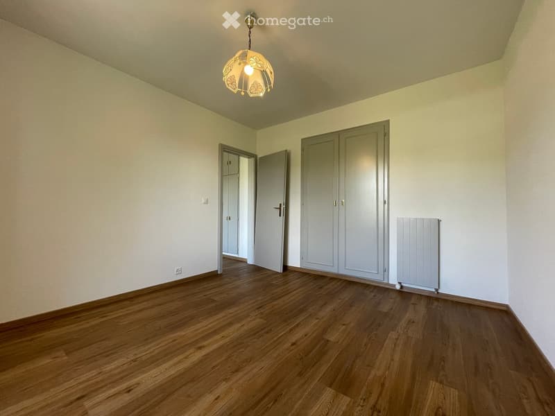 Empty room with wooden flooring, light-colored walls, and a hanging light fixture. Features include a radiator, a closet, and an open doorway leading to another room.