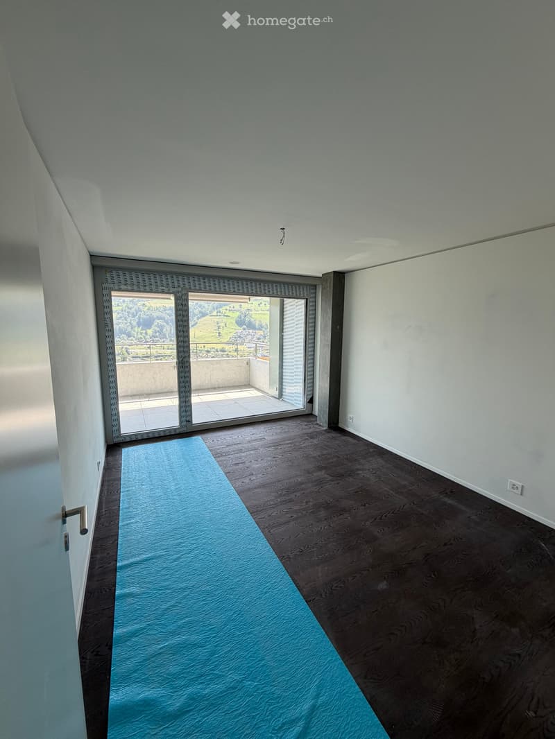 Bright, unfurnished room with wooden flooring, a blue rug, and large sliding glass doors leading to a balcony with scenic views.