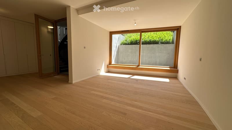 Empty room with light wooden flooring, large window with wooden frame, and a built-in wooden bench seat beneath the window. The room has white walls and a sliding glass door leading to another area.