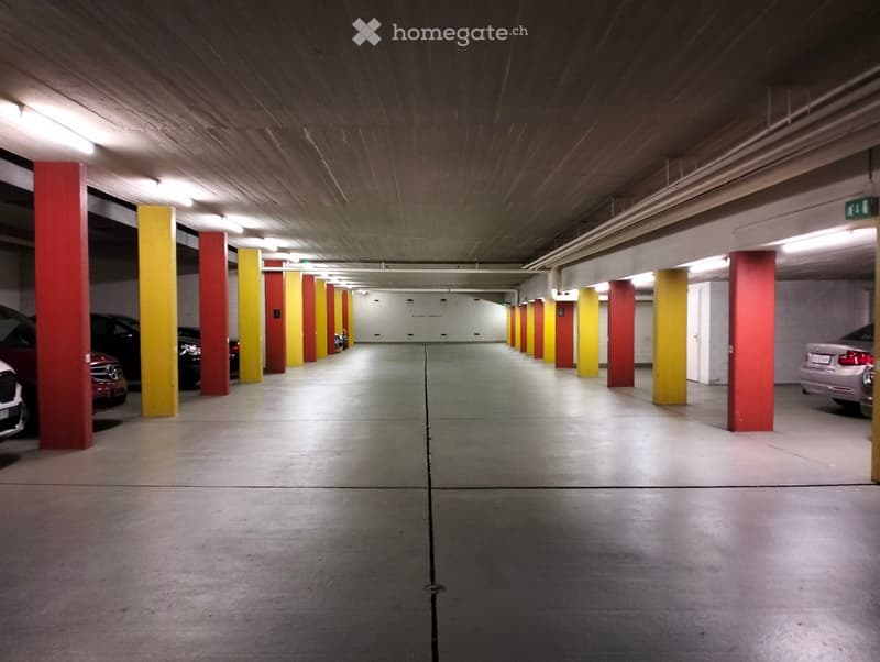 Underground parking area with several parked cars, concrete pillars, and fluorescent lighting.