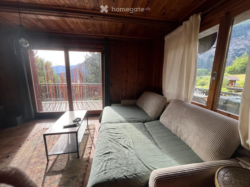 Cozy living room with wooden walls and ceiling, large windows offering a scenic mountain view, a grey sofa, and a coffee table with remote controls.