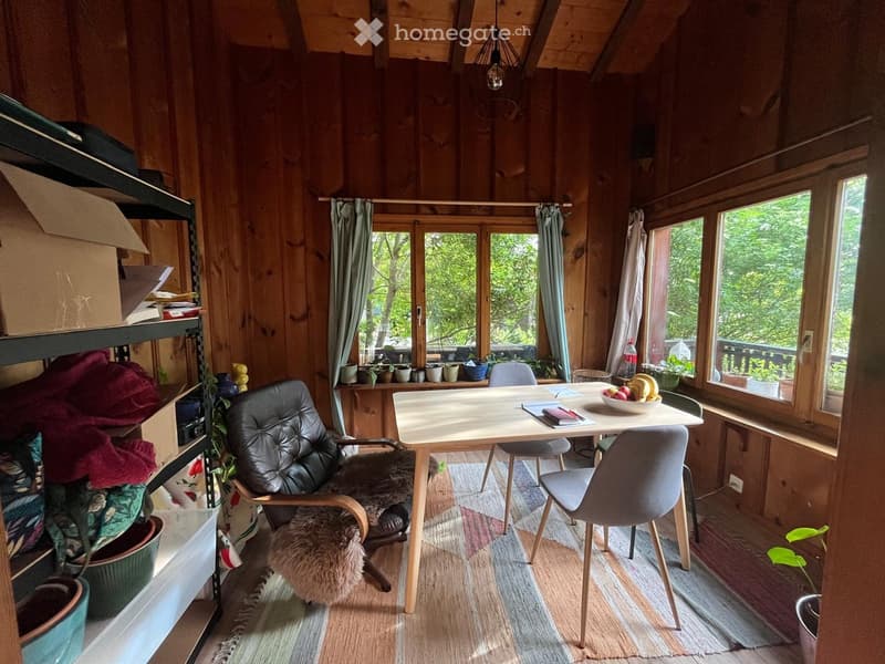 Cozy wooden room with a table, chairs, sofa, shelf, and plants. Large windows offer a view of greenery outside.