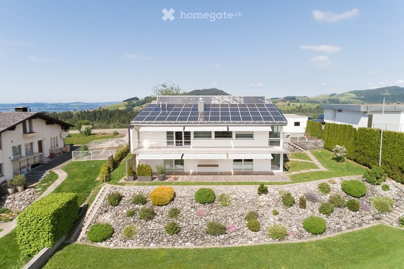 A modern two-story house with a solar panel roof, surrounded by a well-maintained garden with rock beds, shrubs, and a wooden deck area.
