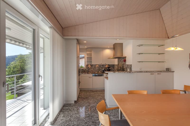 Open-plan kitchen and dining area with white cabinets, granite countertops, a wooden dining table with chairs, and a view of greenery through large windows.