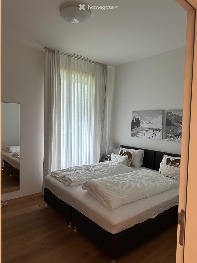 A well-lit bedroom with a large double bed, white bedding, and decorative pillows. The room features wooden flooring, large windows with vertical blinds, a wall-mounted lamp, and artwork depicting a snowy mountain scene.