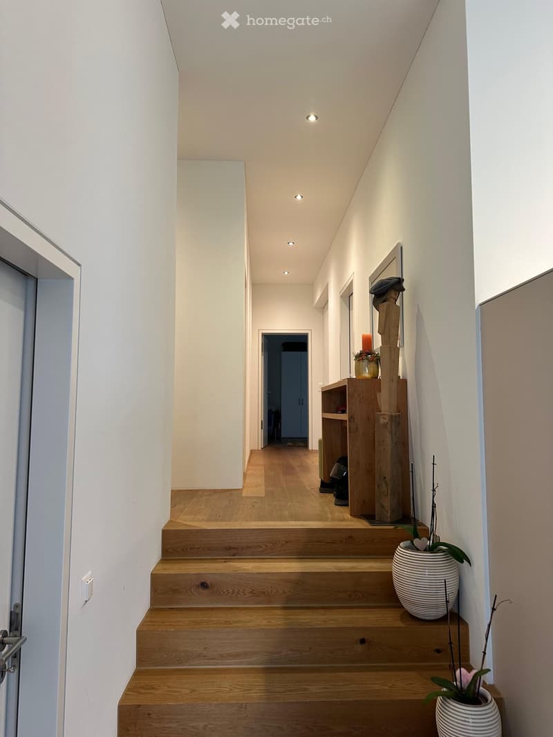 Modern hallway with wooden stairs, white walls, recessed lighting, and decorative items including a vase with flowers and a wooden shelf with shoes and a candle.
