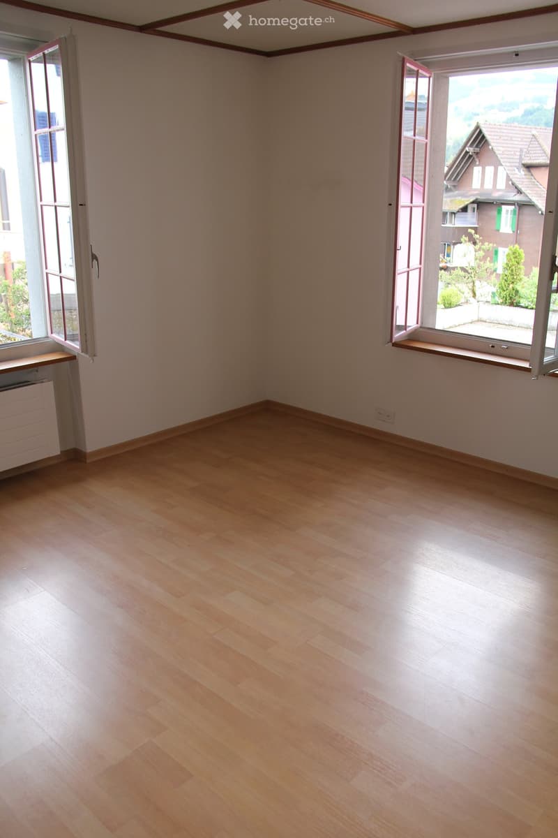 Empty room with light wooden flooring, white walls, and large windows with open shutters, offering a view of the outside area.