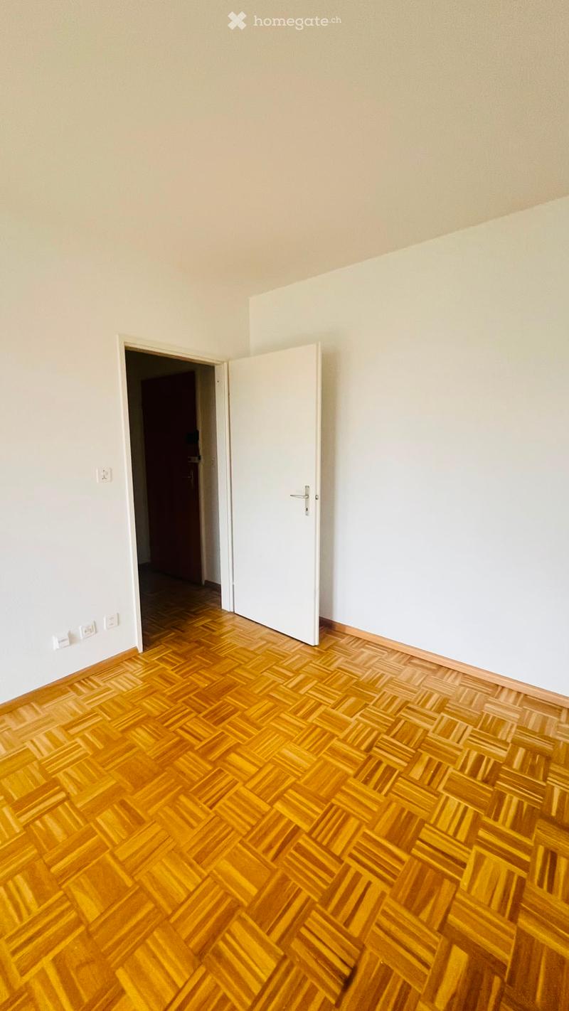 Empty room with wooden herringbone flooring, white walls, and a partially open white door leading to another room.