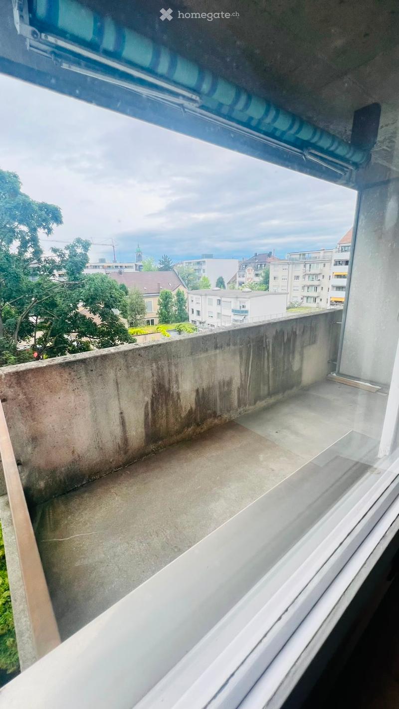 Empty balcony with concrete floor and walls, overlooking a cityscape with buildings and trees.