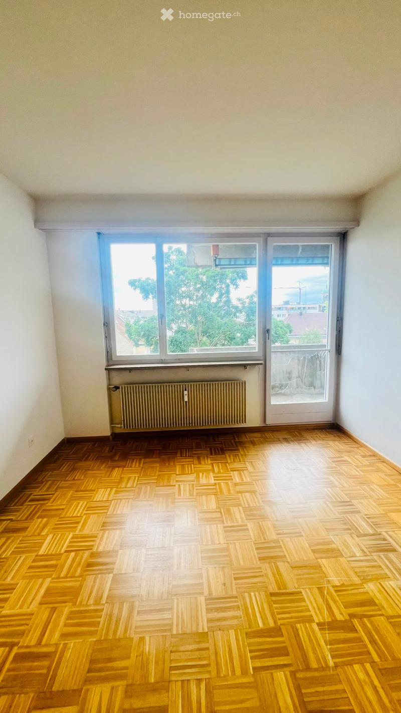 Empty room with wooden parquet flooring, large window with view, radiator, and sliding glass door leading to balcony.
