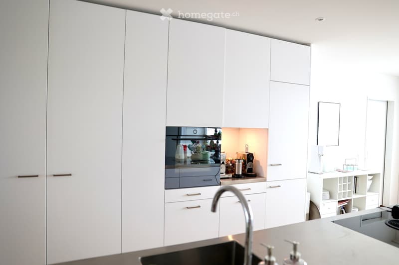 Modern kitchen with white cabinets, stainless steel sink, and various appliances including a fridge, coffee maker, and oven. Shelves hold dishes and other kitchen items.