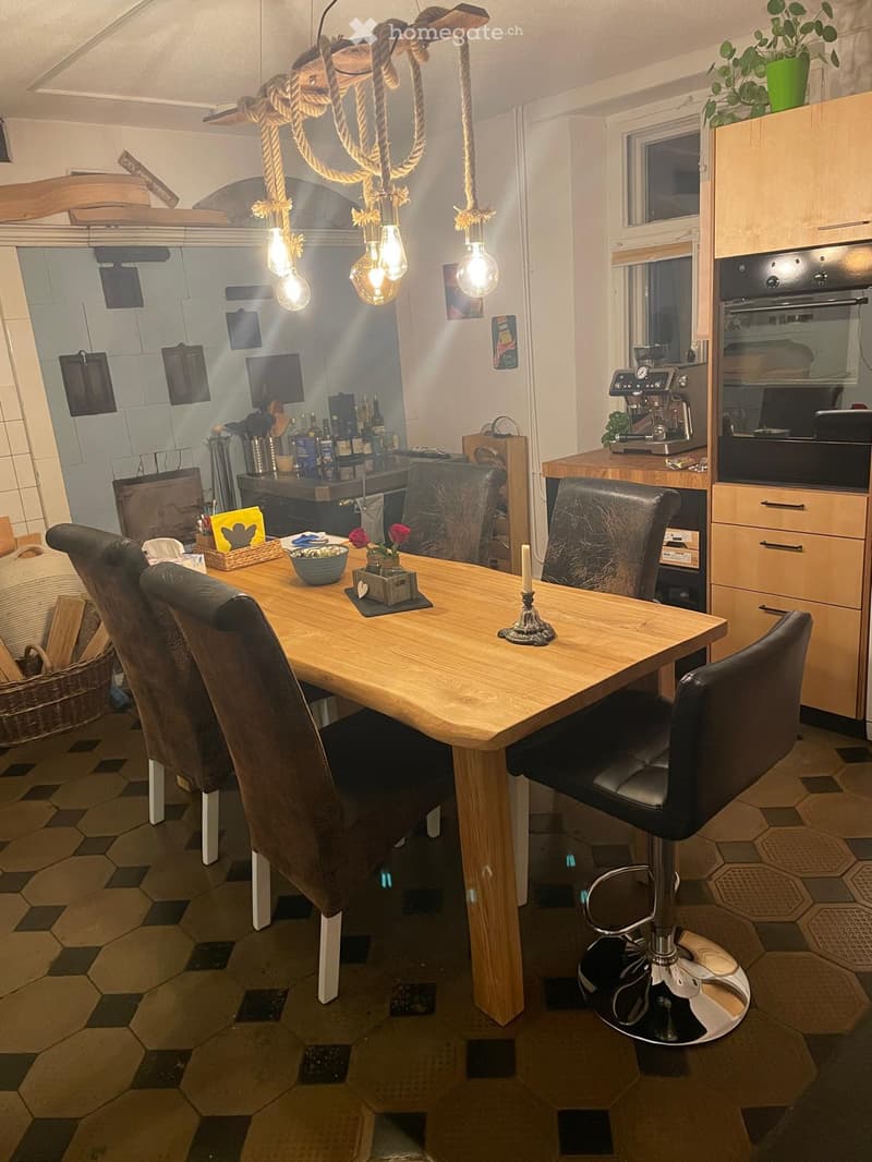 Modern kitchen with a wooden dining table, leather chairs, a bar stool, and various appliances including an oven, refrigerator, and coffee machine. The space features a hanging light fixture and a cozy seating area.