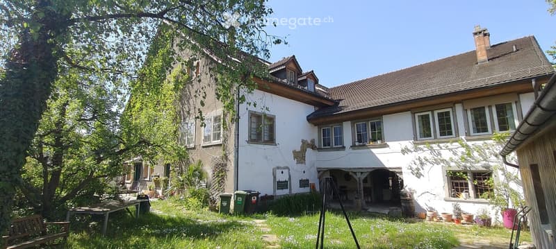 Two-story house with white exterior, multiple windows, and a well-maintained garden area with a bench, table, and potted plants.