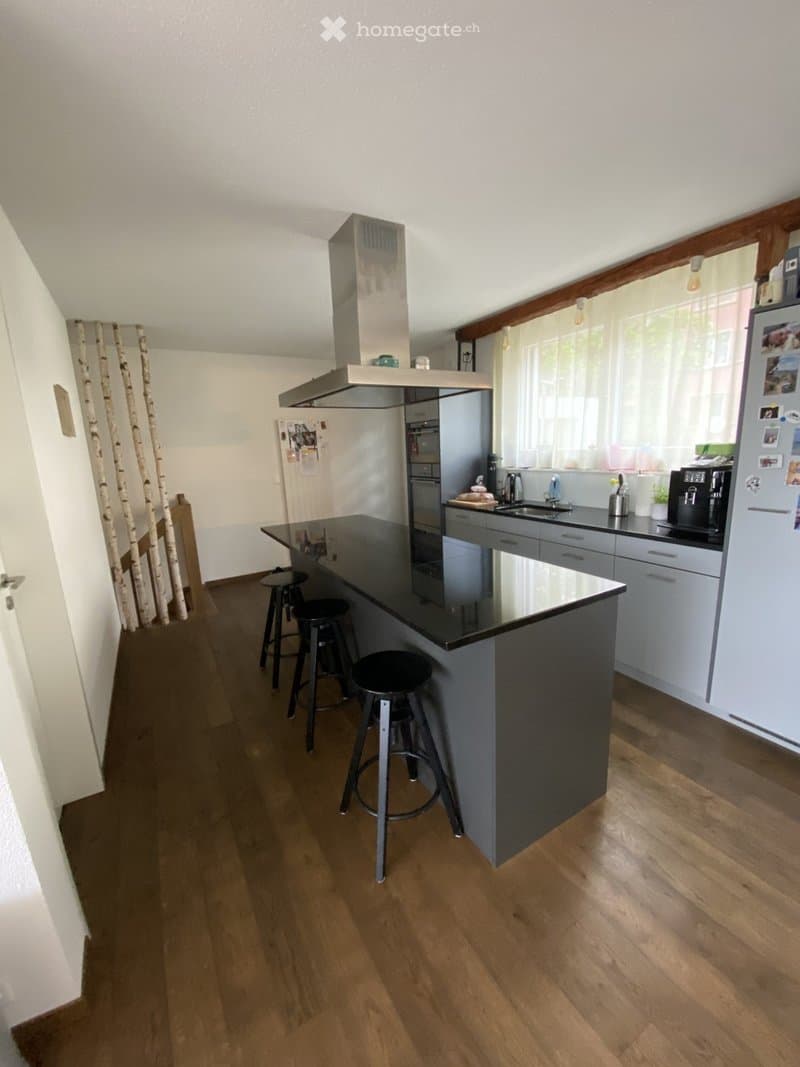 Modern kitchen with a central island, black countertops, grey cabinets, and wooden flooring. Includes a range hood, sink, and various appliances.