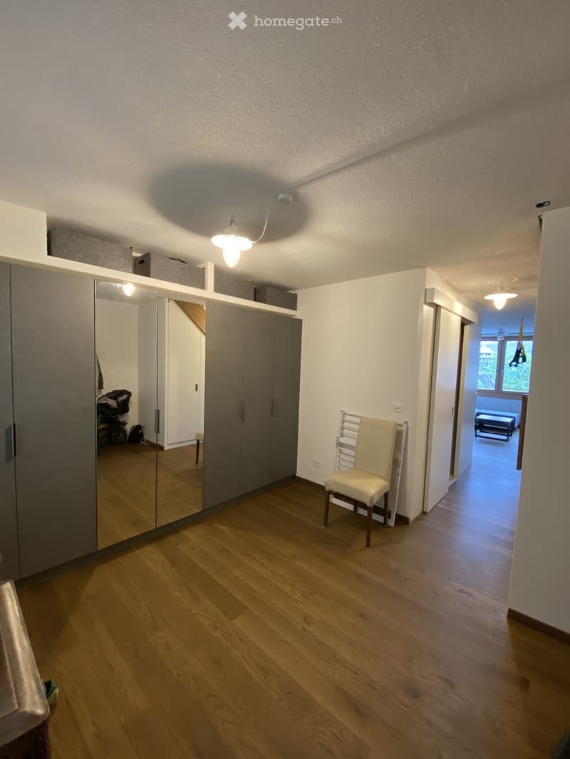 A well-lit hallway with wooden flooring, a large mirrored wardrobe, a single chair, and a visible adjacent room with a sofa and a window view.