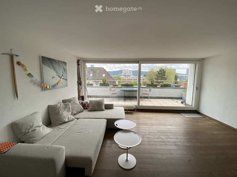 Modern living room with a grey sectional sofa, two white coffee tables, a floor lamp, and a large window opening to a balcony with outdoor seating and a scenic city view.