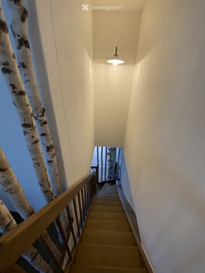 A narrow staircase with wooden steps and a wooden handrail, illuminated by a single hanging light fixture. The walls are painted white, and the staircase leads upward to an open doorway.