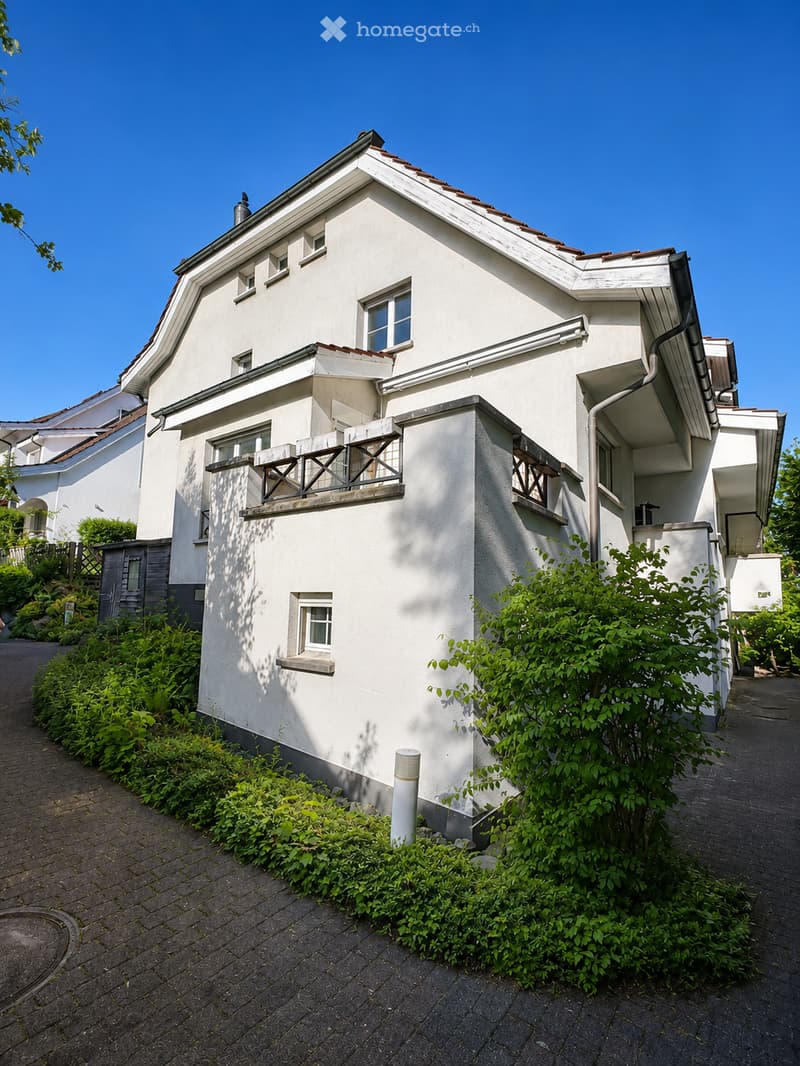 Two-story white house with multiple windows, balconies, and surrounding greenery.