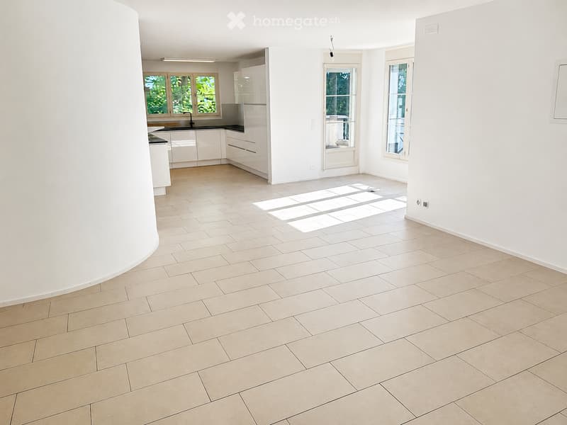 Empty room with tiled floor and white walls, featuring a small kitchen area with cabinets and a sink in the background.