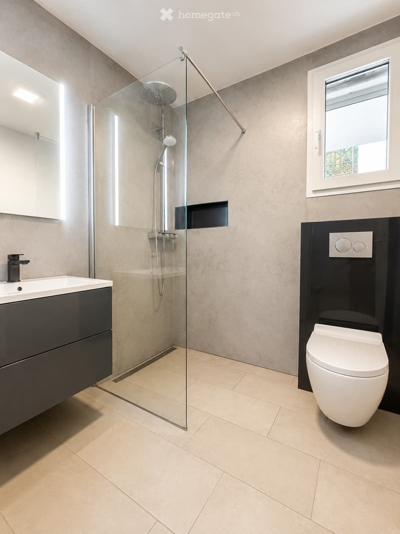 Modern bathroom with beige tiles, a glass-enclosed shower, a white toilet, a black vanity with a sink, and a large mirror.