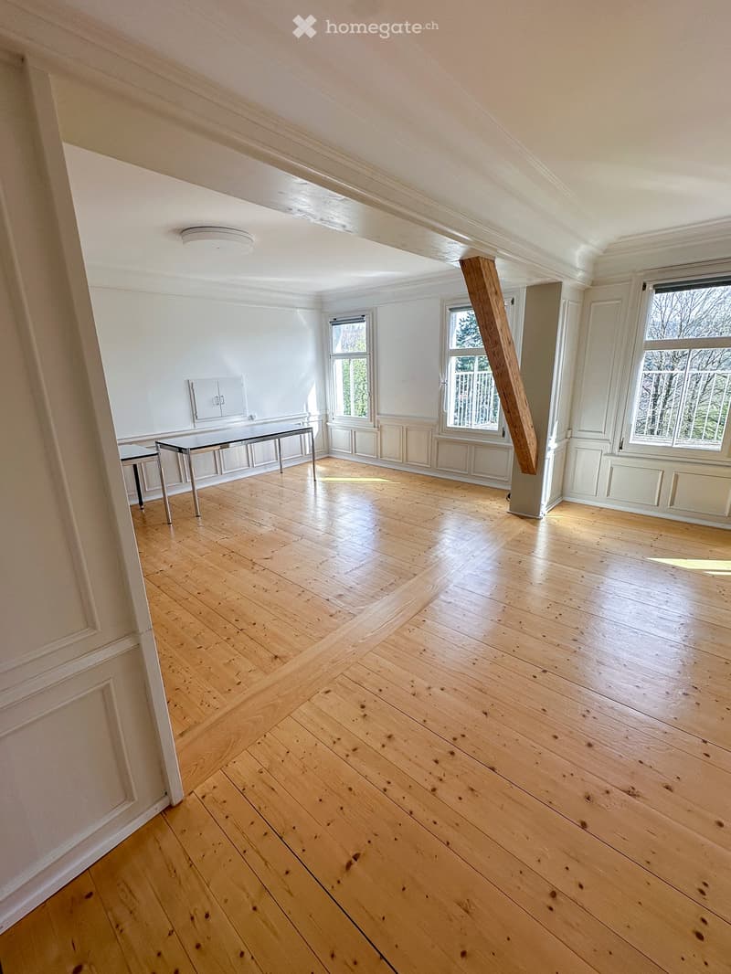 Empty room with light wooden flooring, white walls, and large windows allowing natural light. The room features a modern, minimalist design with a visible structural wooden beam and a small table near the entrance.