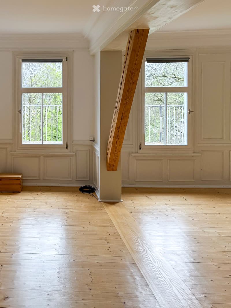 Empty room with wooden flooring, white walls, large windows with metal railings, and a wooden chest in the corner.
