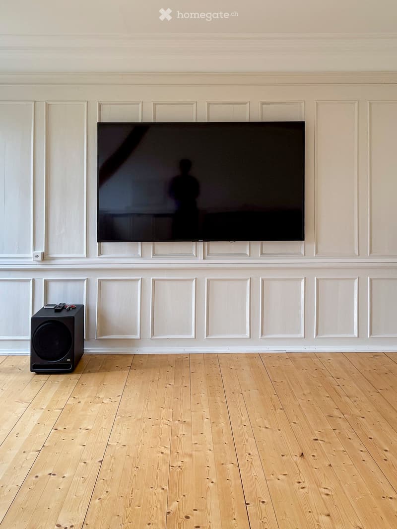 A modern living room with a large flat-screen TV mounted on a white paneled wall, a black speaker on the floor, and a pair of remote controls on top of the speaker. The room features light wooden flooring and white walls with decorative paneling.