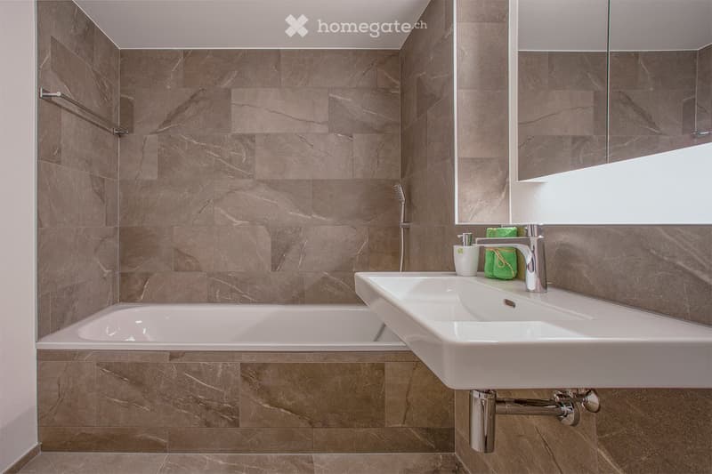 Modern bathroom with a bathtub, double sink, and large mirror. Brown marble tiles line the walls and floor, and a towel rack is mounted on the wall.