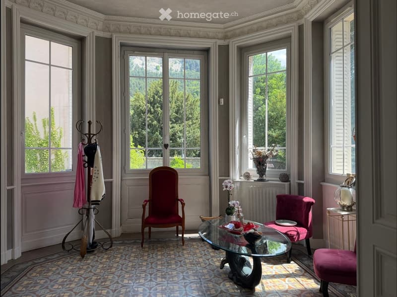 Elegant living room with large windows, ornate ceiling, patterned tile floor, red upholstered chairs, glass coffee table, decorative vase, and coat rack.