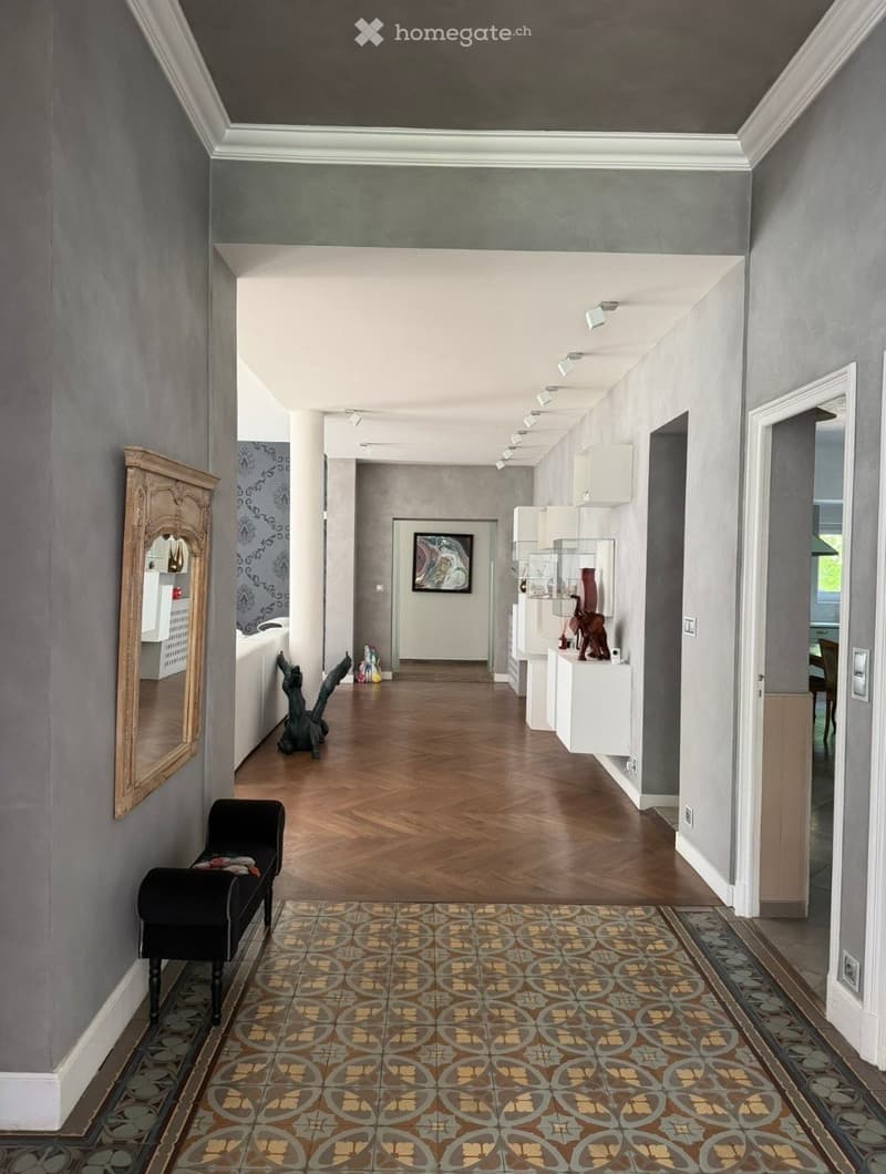 Elegant hallway with wooden flooring, grey walls, white ceiling moldings, and modern lighting. Features a patterned rug, a small black bench, and decorative items including a mirror, artwork, and sculptures.