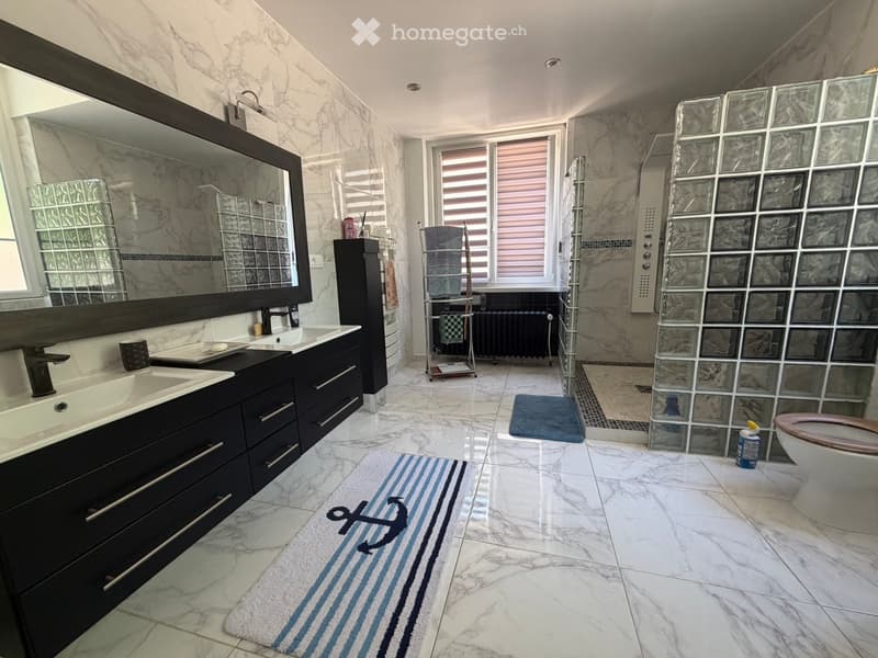 Modern bathroom with marble tiles, double sink vanity, large mirror, shower with glass blocks, and a towel rack. Features a blue and white striped rug with an anchor symbol.